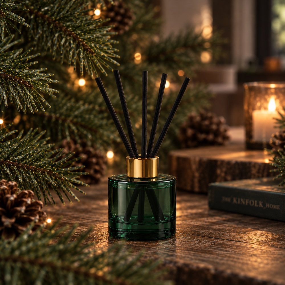 A green glass reed diffuser bottle with black reeds and a gold cap, styled on a wooden table with Christmas pine branches, pinecones, and a warm candle in the background.